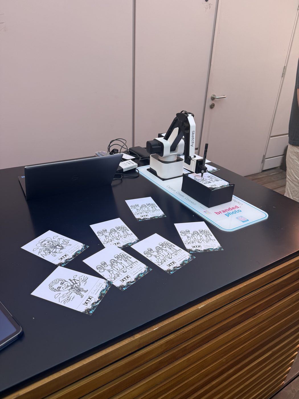 A sketch robot arm drawing on branded cards at a corporate summer party, with finished portraits laid out on the table.
