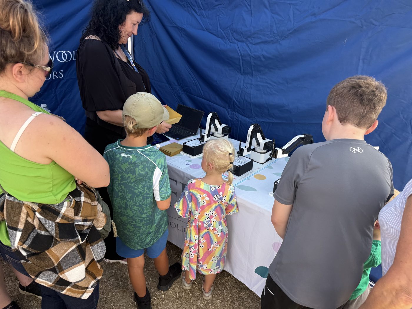 Families and children gathered around three sketch robots on an exhibitor stand at an outdoor county show.