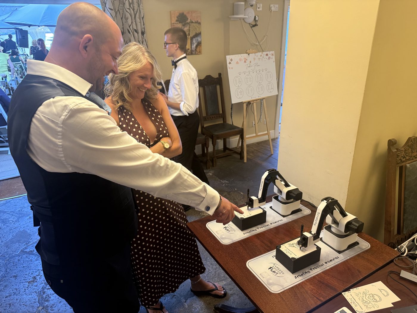 Wedding guests watching two robot arms draw portraits on a table during a reception.